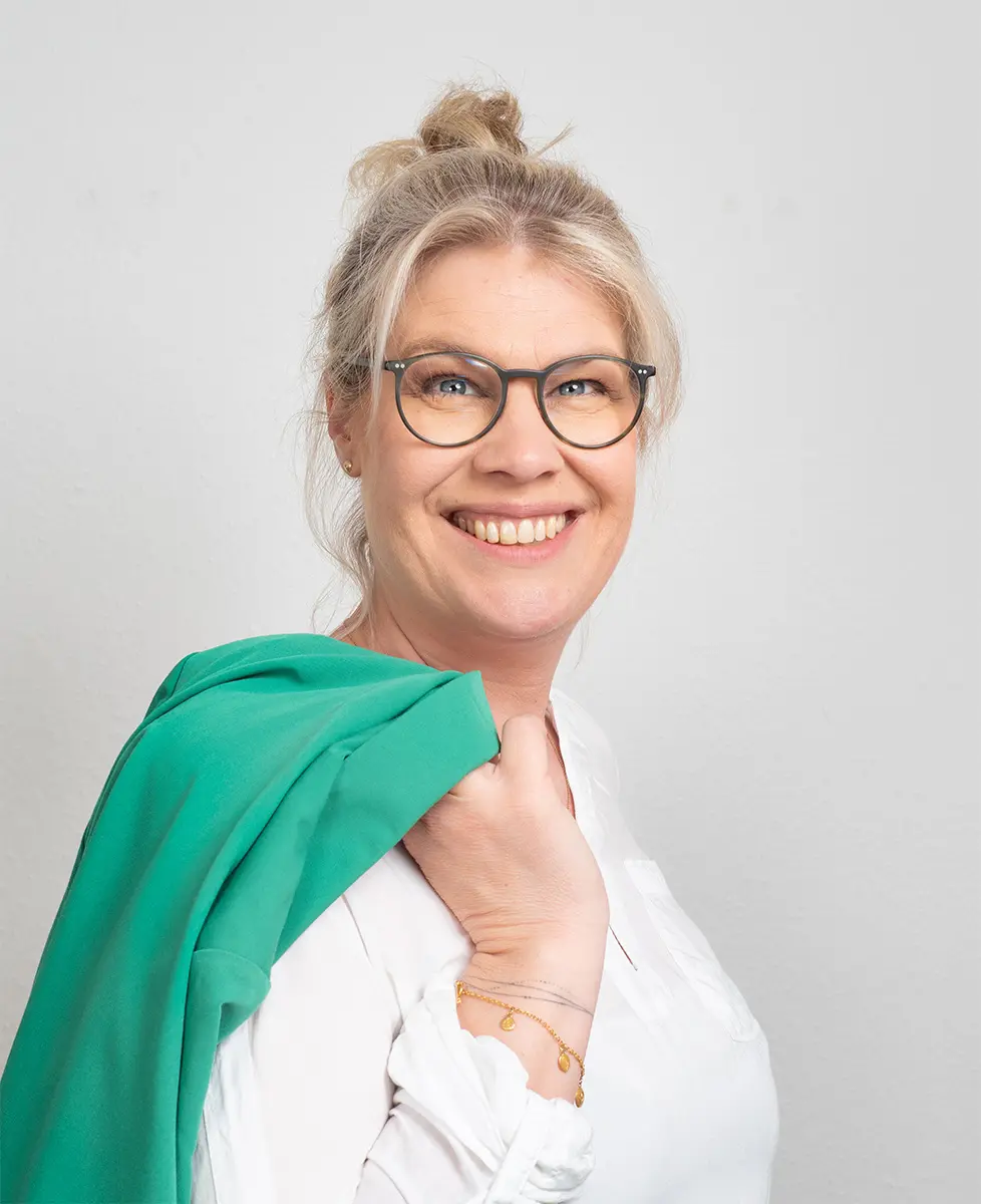 Frau mit blonden Haaren und Brille, die ein grünes Kleidungsstück über der Schulter trägt.