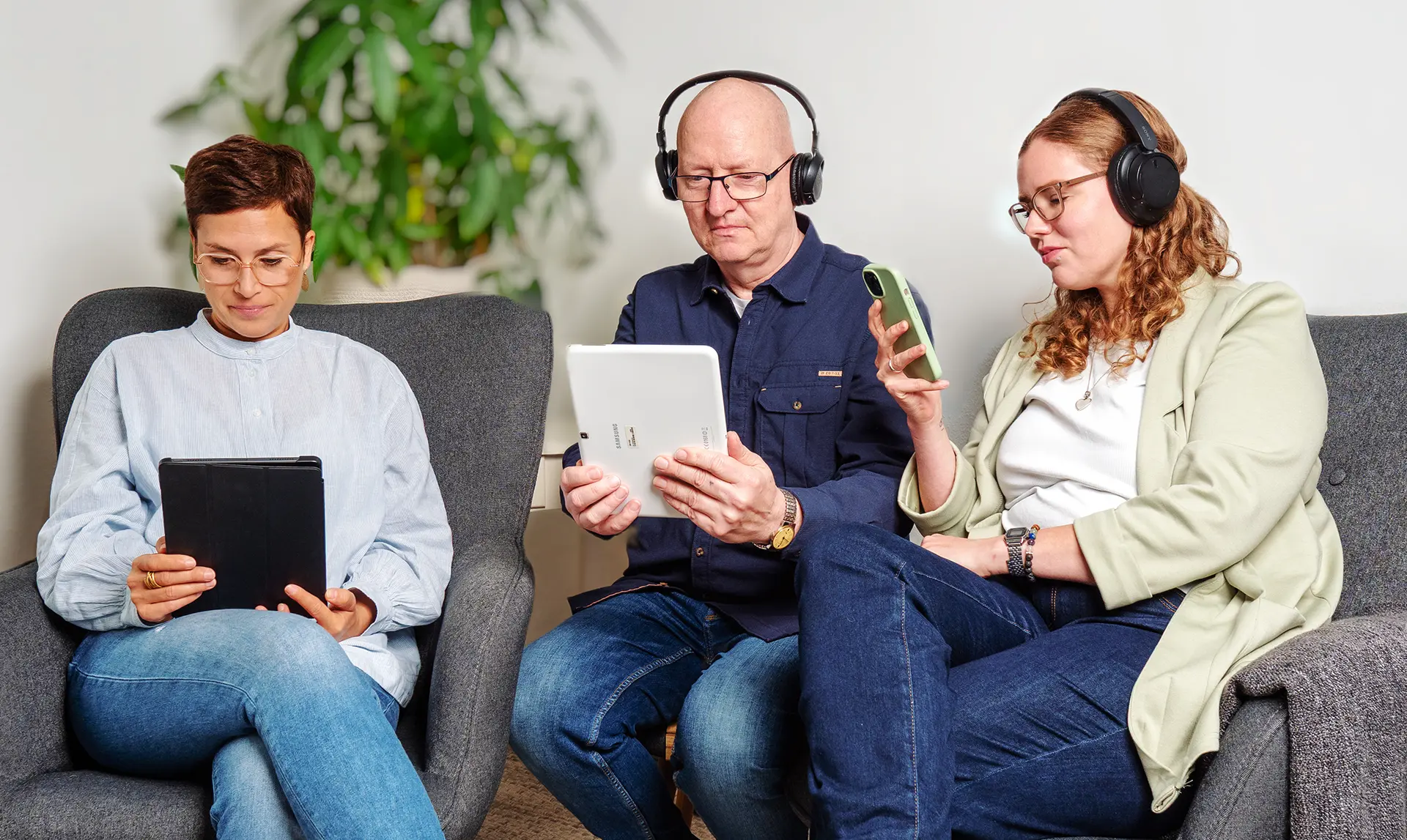 Drei Personen sitzen auf einem Sofa, tragen Kopfhörer und nutzen Tablets und Smartphones.