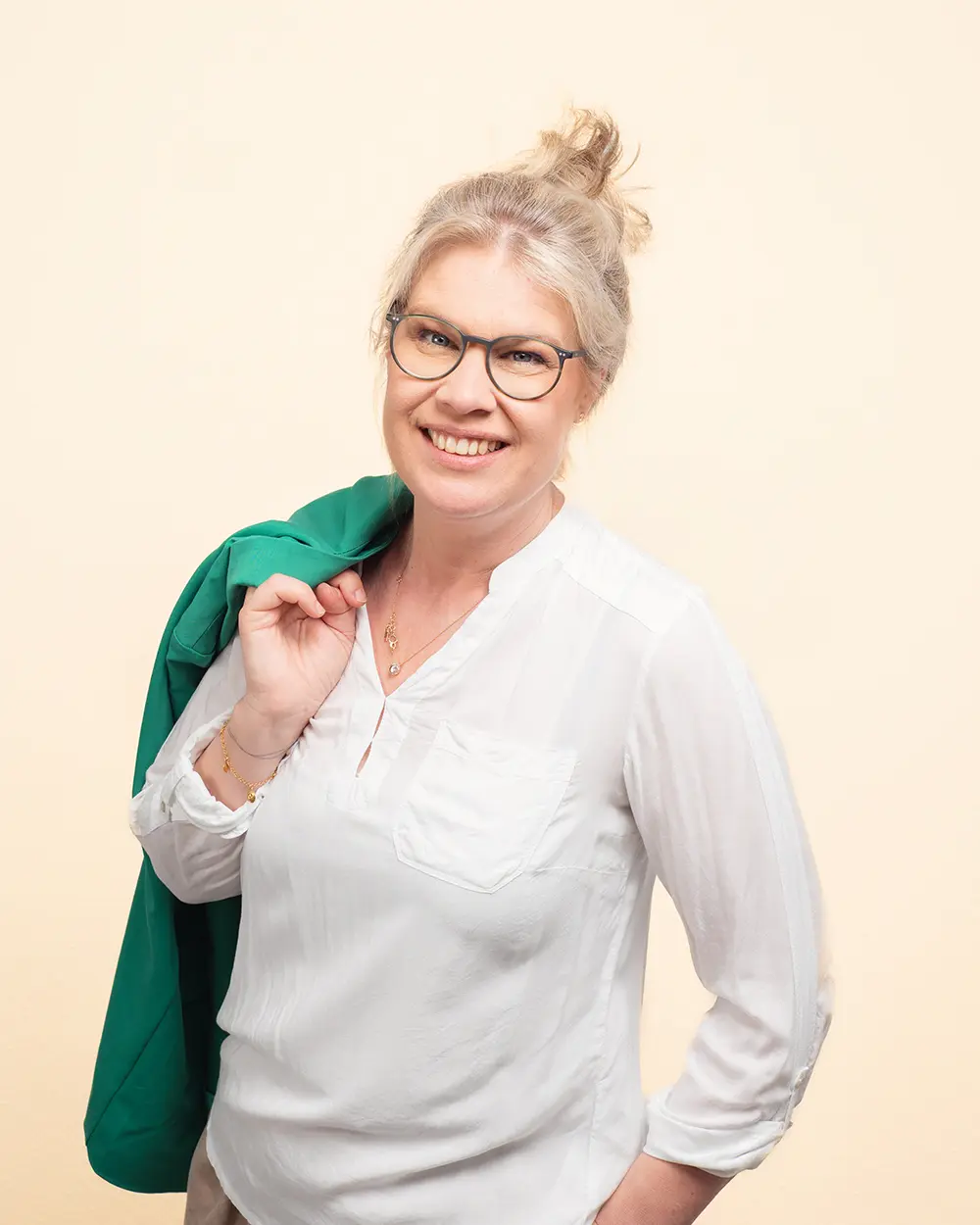 Frau mit blonden Haaren, Brille und einer grünen Jacke auf der Schulter, lächelt in die Kamera.